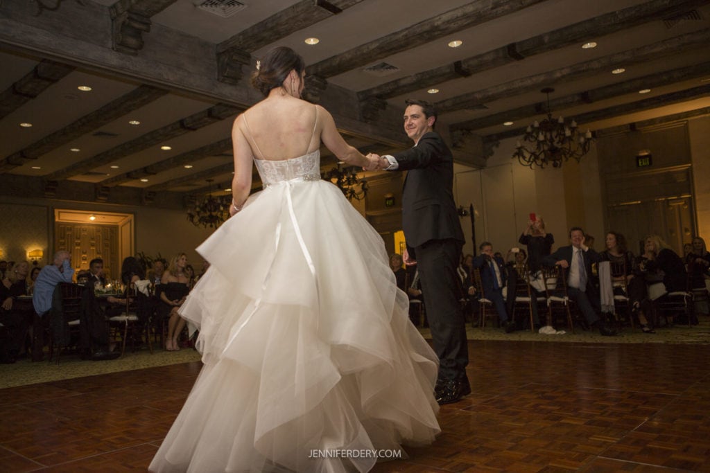 A bride in a strapless white wedding gown and a groom in a black suit share a dance on a wooden floor in the dimly lit venue of Founders Chapel. Guests seated around the room watch the couple, and an ornate ceiling with exposed beams adds to the elegant setting.