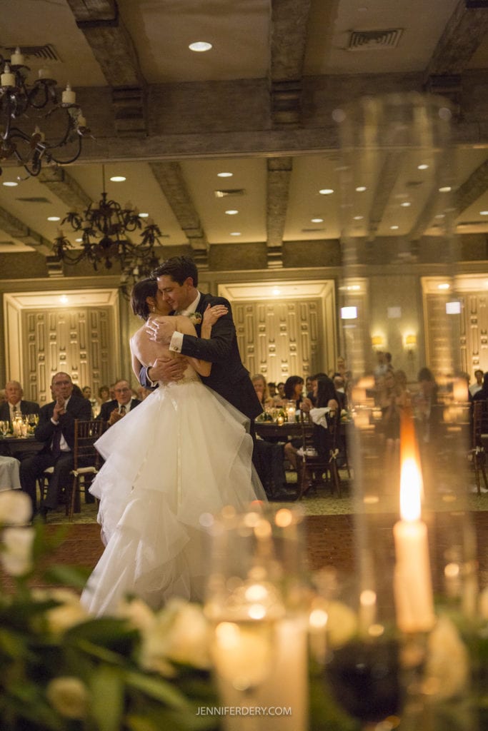 A bride and groom share their first dance in a warmly lit, elegant ballroom at Rancho Bernardo inn. The bride wears a flowing white gown, and the groom is in a dark suit. Guests are seated at round tables in the background, while lit candles add a cozy ambiance to the foreground—perfect for Founders Chapel wedding photos.