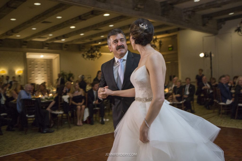 A man in a suit and a woman in a white wedding dress share a dance in the warmly lit reception hall of Founders Chapel, filled with seated guests. The bride's dress has a full skirt, and she has her hair styled in an elegant updo, captured perfectly in their wedding photos.