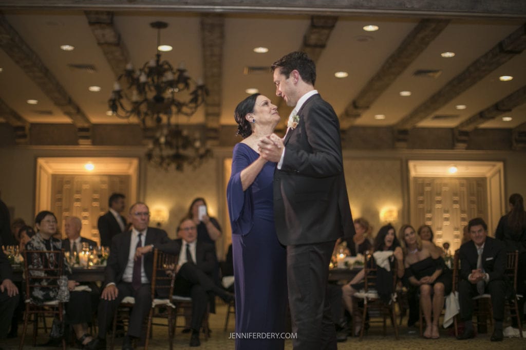 A groom in a black suit dances with an older woman in a blue dress at Founders Chapel Wedding Photos. Guests seated at tables in the dimly lit room attentively watch their graceful movements. The atmosphere is elegant, with chandeliers and decorative wall panels adding to the charm.