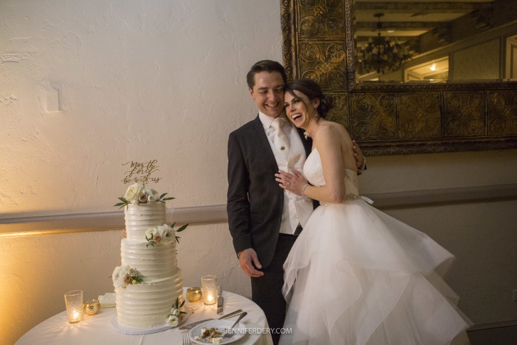 A bride and groom stand closely together, smiling and laughing, next to a three-tiered white wedding cake adorned with flowers. The cake topper reads "Mr & Mrs." The warmly lit room with an ornate mirror behind them adds charm to their Founders Chapel wedding photos.