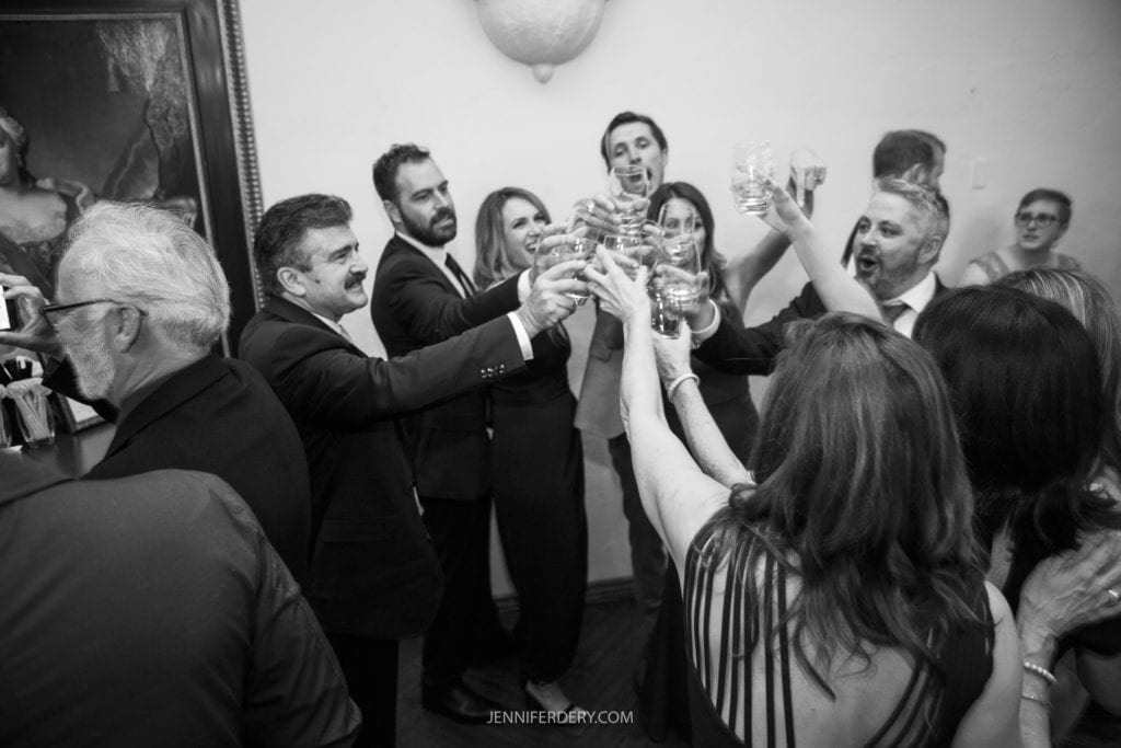 A group of people raising their glasses in a celebratory toast at a gathering. They are dressed in formal attire, with smiles and laughter visible. The setting is indoors, resembling the warmth of Founders Chapel wedding photos, with framed artwork hanging on the wall in the background.