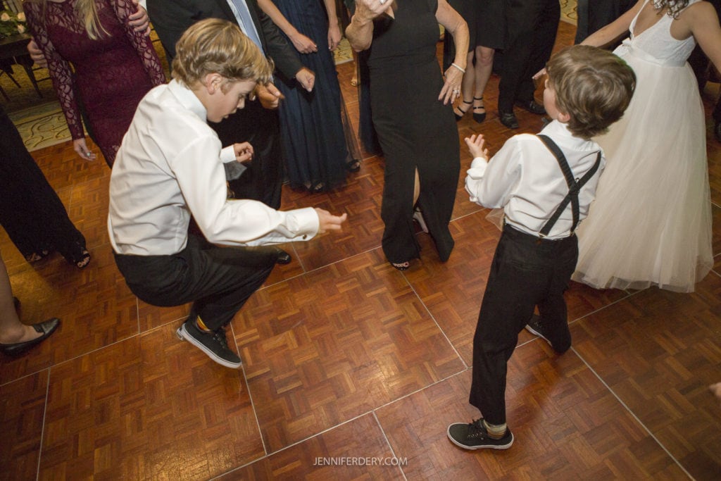 Two young boys energetically dance on a wooden floor at a Founders Chapel wedding. One boy in a white shirt, black pants, and sneakers is bending down, while the other boy in a white shirt with suspenders is smiling. Elegantly dressed guests surround them, creating the perfect wedding photo moment.