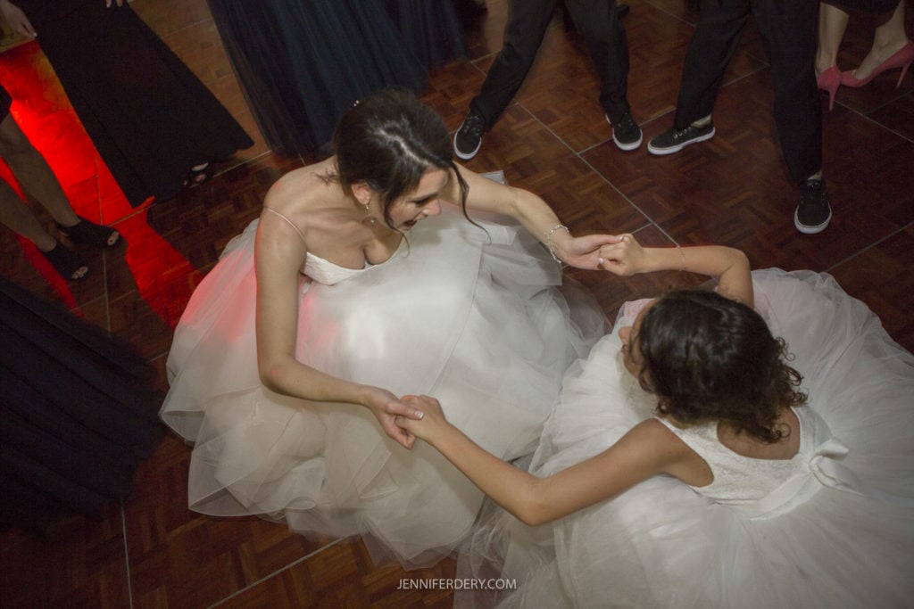 Two women in white, tulle dresses are holding hands and smiling at each other while dancing on a wooden floor. The photo, part of their Founders Chapel wedding photos, is taken from above. One is standing and the other kneeling. Other people in formal attire are seen dancing around them.