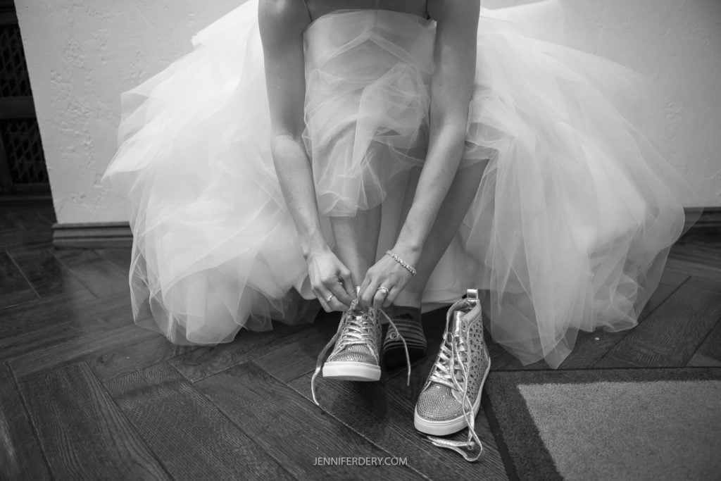A bride in a voluminous tulle wedding dress bends down to tie the laces of her sparkly high-top sneakers. The black and white photo, taken at Founders Chapel, captures this unique moment, blending elegance and casual style. The wooden floor beneath adds a rustic touch to the scene.