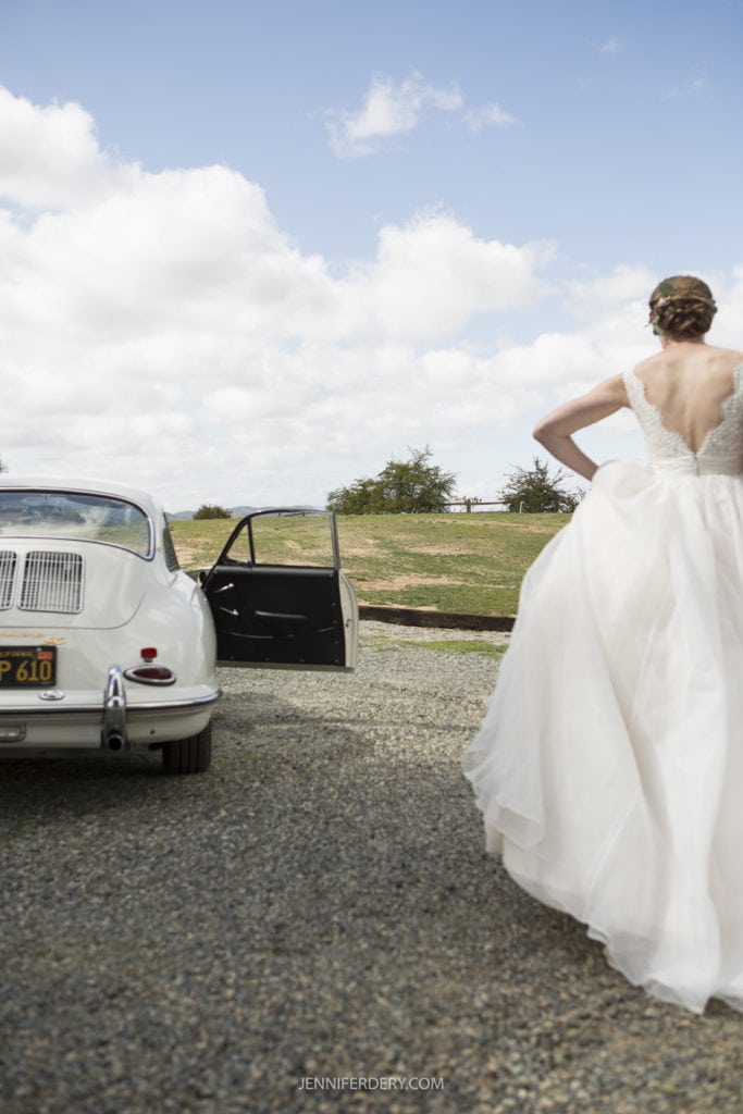 A bride in a white wedding dress stands with her back to the camera, facing an open door of a vintage white car parked on a gravel path. The rustic wedding scene is set outdoors under a partly cloudy sky, with grassy fields and sparse trees in the background.