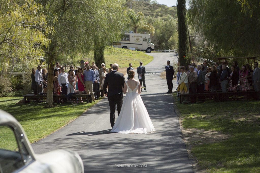 A bride and groom walk hand-in-hand down an outdoor aisle lined with guests on either side. Tall trees and greenery surround the area, enhancing the rustic wedding setting, and an old-fashioned school bus is parked in the background. The scene is bright and sunny, creating a romantic atmosphere.