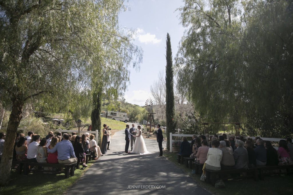 An outdoor wedding ceremony with the couple standing in the center under a tree, exchanging vows. Guests are seated on wooden benches on both sides of the aisle, surrounded by lush greenery and a sunny sky. A windmill and a rustic fence add to the charm of this picturesque rustic wedding setting.