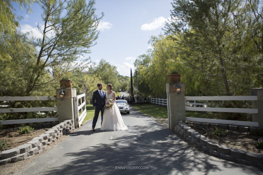 A newlywed couple walks down a scenic driveway at their rustic wedding, with trees lining both sides and a vintage car visible in the background. They share a joyful moment on a sunny day, holding hands and gazing at each other. The word "JENNIFRIEDERY.COM" is at the bottom center.
