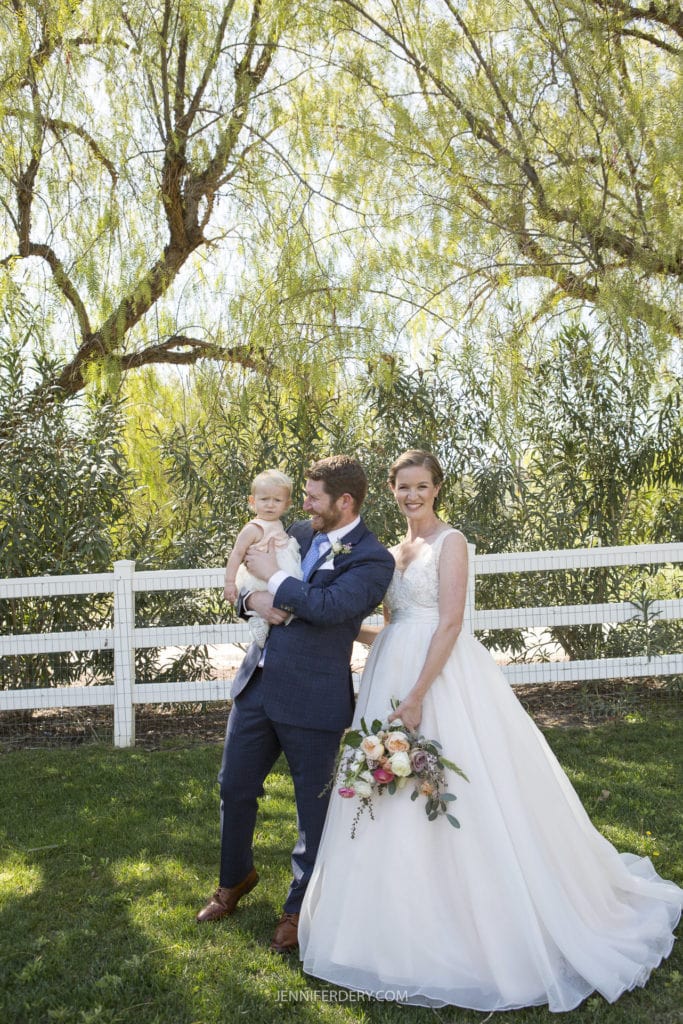 A bride and groom stand together outdoors in front of a white fence and greenery at their rustic wedding. The bride, in a white gown holding a bouquet of flowers, smiles while the groom, in a dark suit and bowtie, holds a young child in his arms. All appear happy and relaxed.