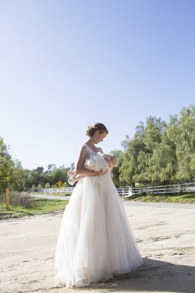 A bride in a flowing white wedding dress cradles a baby in her arms while standing on a sandy pathway. The rustic wedding background features green trees, a white fence, and a clear blue sky.