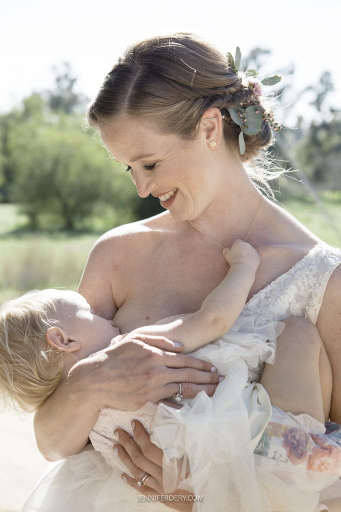 A woman in a sleeveless white dress is breastfeeding an infant dressed in a white outfit. The woman has a small bouquet of flowers in her hair and is smiling while looking down at the child. They are outside with greenery in the background, capturing a serene moment amidst the rustic wedding setting.
