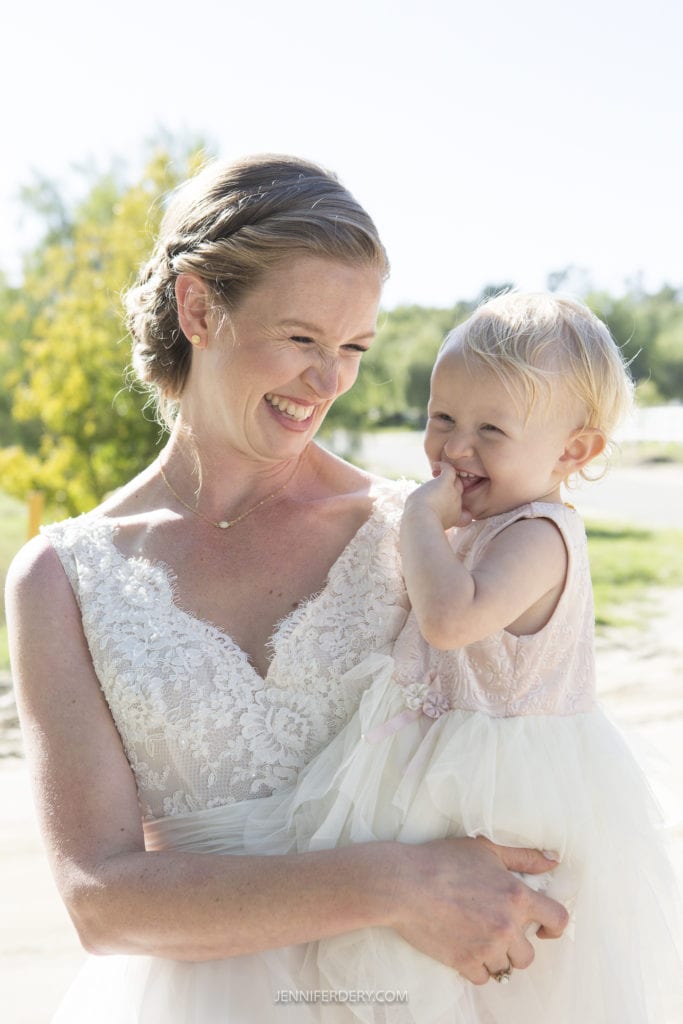 A joyful woman in a white lace wedding dress holds a young child in her arms at a rustic wedding. They are outdoors on a sunny day, both smiling and laughing. The child, wearing a light pink dress, touches their face with a finger. Trees and greenery are visible in the background.