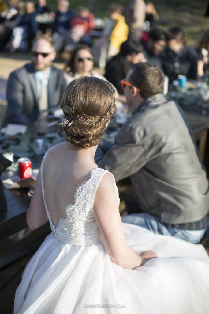 A bride in a white lace dress with her hair braided and adorned with flowers is sitting at a wooden table outdoors, capturing the essence of a rustic wedding. Her back is to the camera. People around the table are engaged in conversation, and a man in a leather jacket is seated next to her.