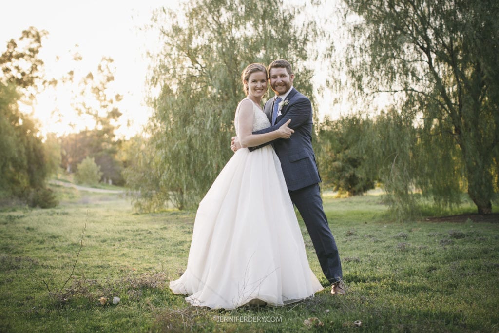 A bride and groom stand embracing each other on a grassy field with trees in the background. The bride is wearing a white dress and the groom is in a suit. The sun is setting, casting a warm glow over their rustic wedding. The mood is romantic and joyful.