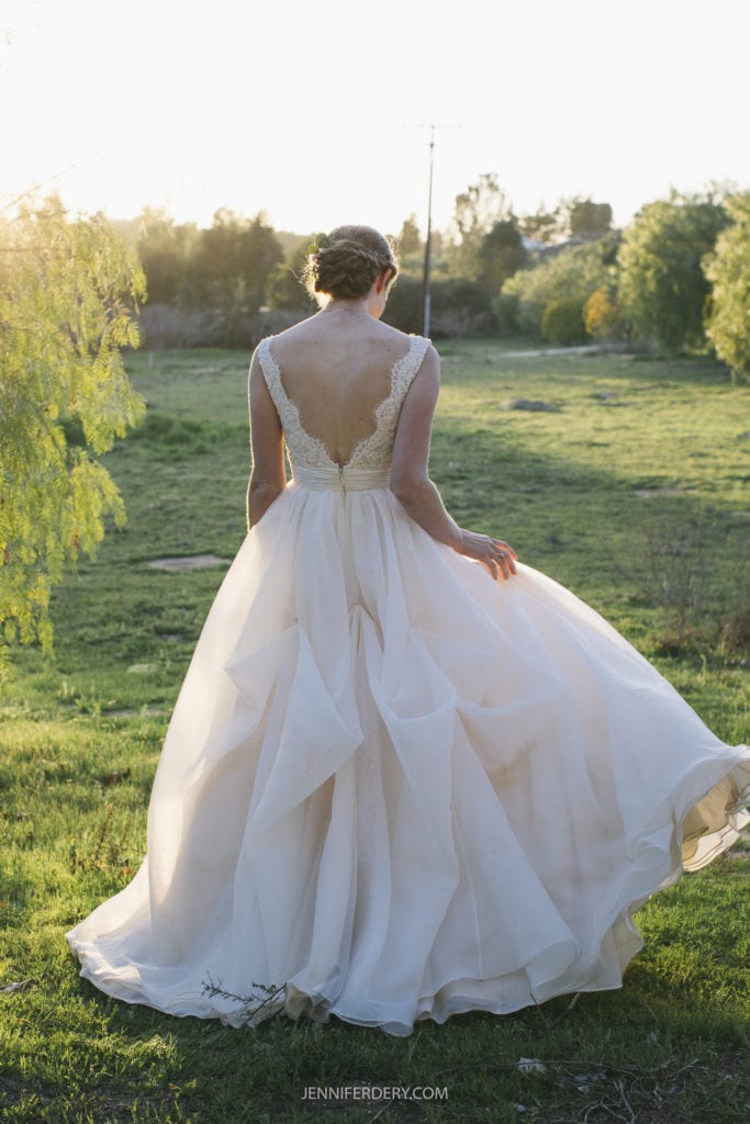 A bride stands outdoors at sunset, facing away from the camera during her rustic wedding. She wears a flowing white wedding dress with a deep V-back and lace detailing. Her hair is in an updo, and she gently holds the skirt of her dress, creating a graceful, elegant pose against a lush, green field with trees.