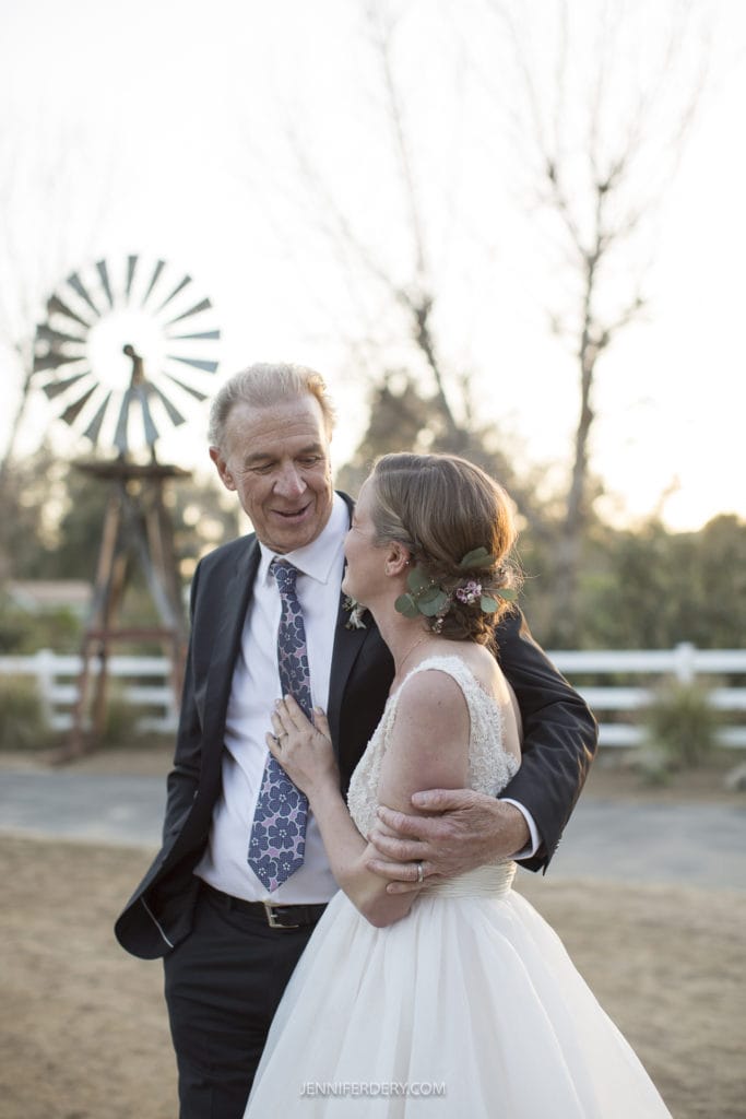 A bride in a white wedding dress shares a joyful moment with an older man in a suit and tie during their rustic wedding. They're outdoors with a blurred background featuring a windmill and wooden fence. The bride, adorned with flowers in her hair, looks at the man and smiles warmly.