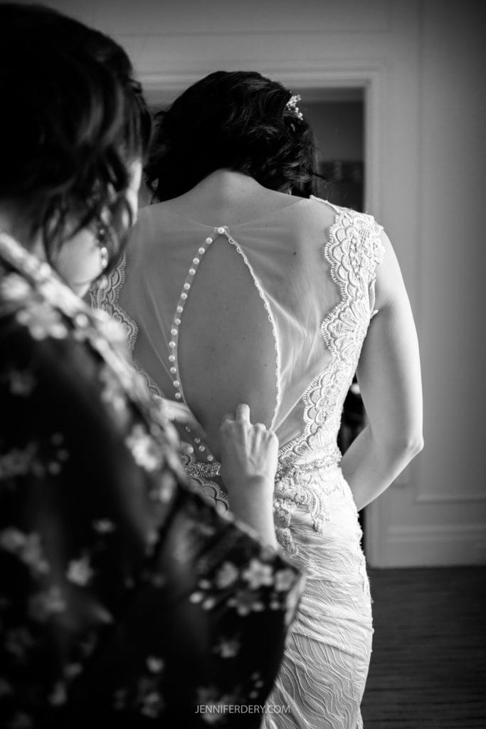 A black-and-white photo captures a tender moment at a Japanese Friendship Garden wedding. The focus is on the back of the bride's dress, featuring sheer fabric with decorative buttons. Another person, partially visible in a patterned outfit, assists her with delicate care.