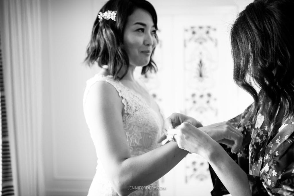 A black-and-white photo of a bride wearing a lace dress, being helped by another woman. The bride has shoulder-length hair adorned with a hair clip and smiles slightly as the other woman, partially out of view, adjusts her bracelet at their Japanese Friendship Garden wedding.