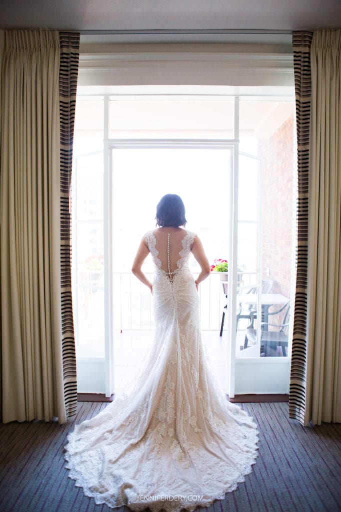 A bride stands facing an open balcony door, framed by beige curtains. She wears a white, lace wedding dress with a long train that cascades to the floor. The back of the dress features an intricate lace pattern and a row of buttons. Natural light filters in, evoking the peace of a Japanese Friendship Garden Wedding.