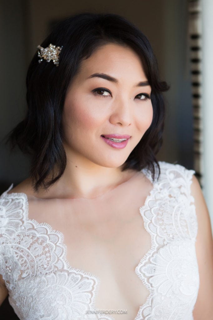 A woman with shoulder-length black hair, adorned with a floral hairpin, smiles softly at her Japanese Friendship Garden wedding. She is wearing a white lace dress with an elaborate design and a plunging neckline. The background is softly focused.
