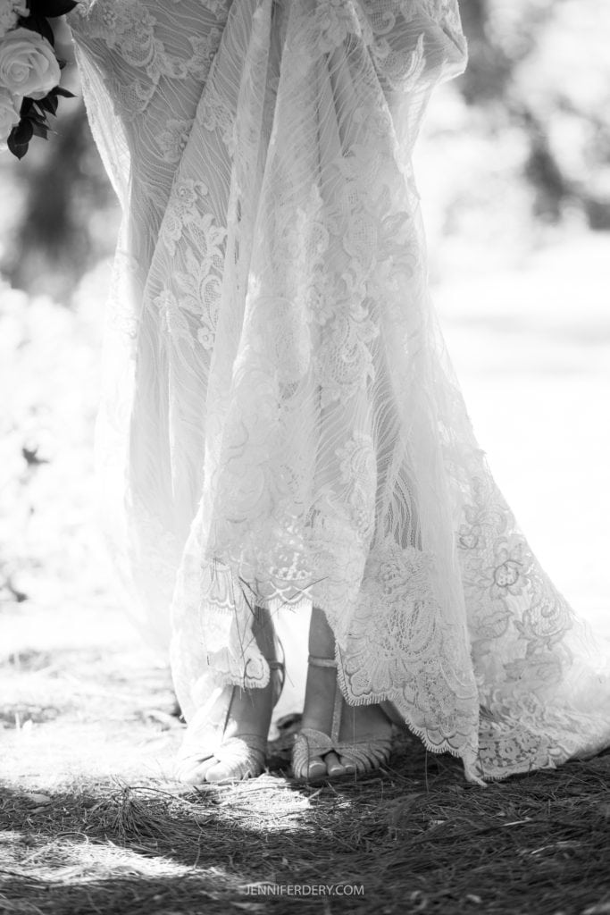 Black and white photo of a bride lifting her lace gown slightly, revealing her feet in strappy high-heeled sandals. The intricate lace details of the dress are prominent, with delicate floral patterns. Captured at a Japanese Friendship Garden Wedding, the background is blurred to focus on the bridal attire.