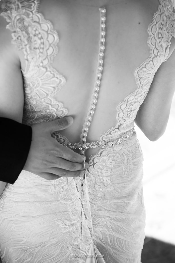 A black and white photo of a bride in a lace wedding dress, seen from behind, at a Japanese Friendship Garden wedding. The dress features an intricate lace design and a row of buttons along the back. A hand, presumably of a groom or attendant, is gently touching her back near the waist.