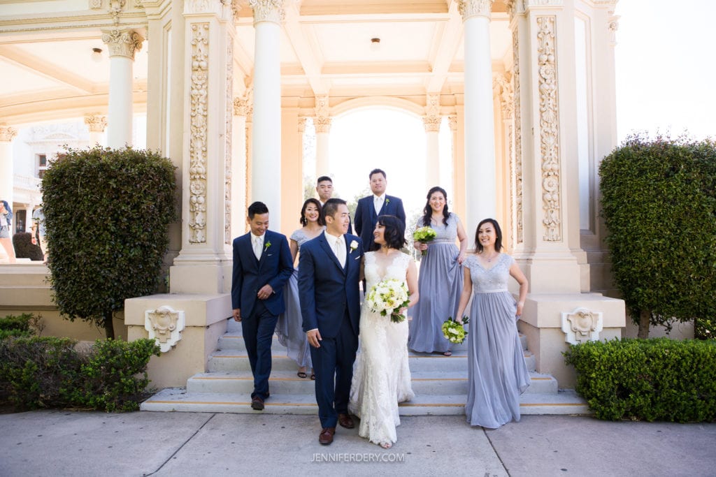 A bride and groom, dressed in traditional wedding attire, walk down steps outside the elegant Japanese Friendship Garden. The groom is in a navy suit, and the bride holds a bouquet. Six attendants follow, dressed in matching grey outfits, smiling and chatting at their serene garden wedding.