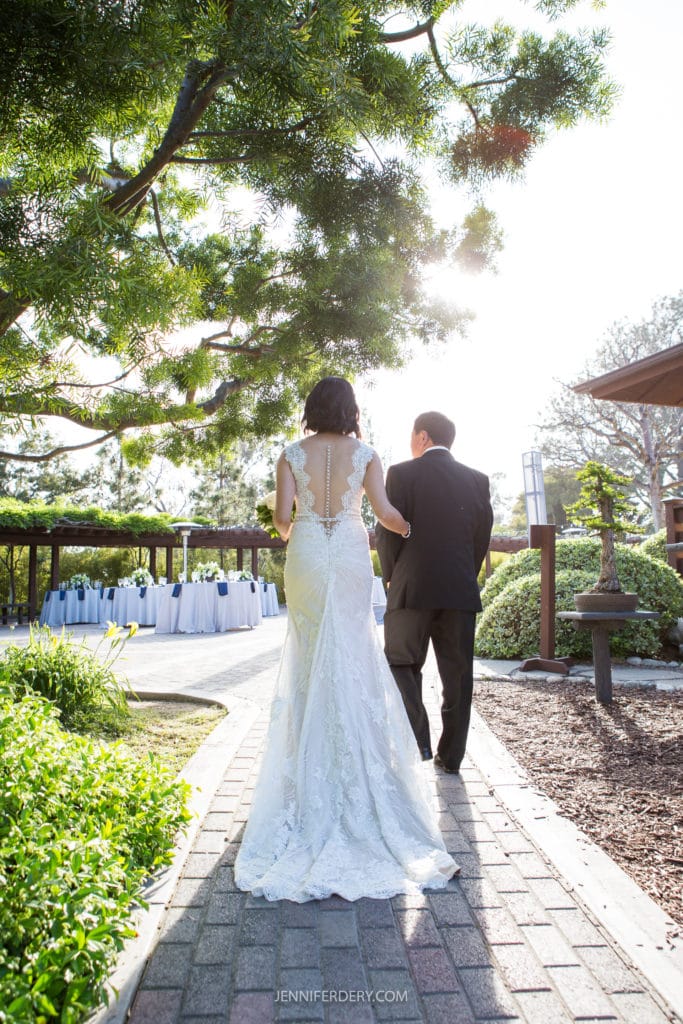A bride in a lace wedding gown walks arm-in-arm with a man in a black suit down a brick path at the Japanese Friendship Garden Wedding towards an outdoor reception area. The sun is shining through the trees, and tables with white tablecloths are set up under a gazebo.