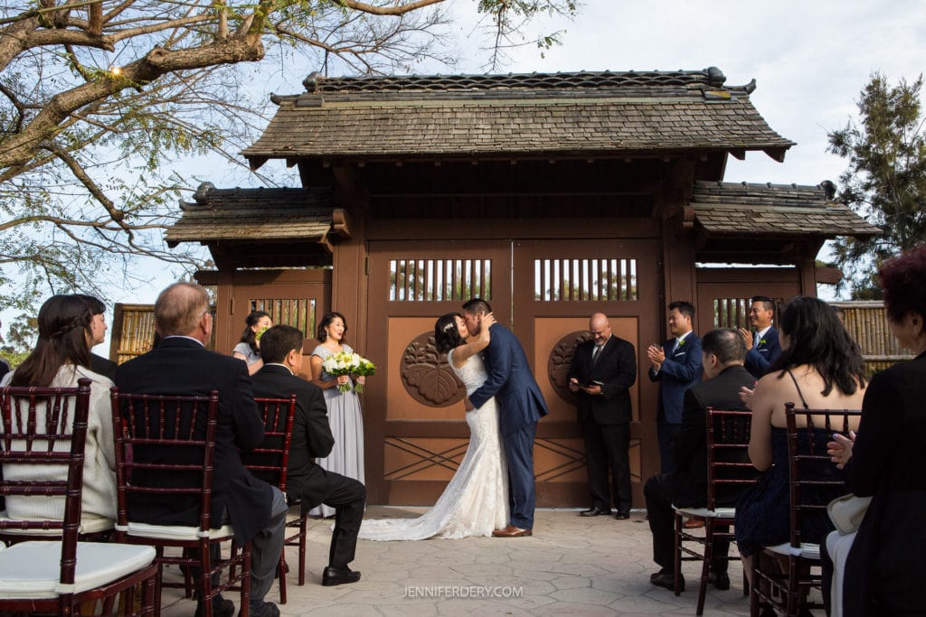 A bride and groom share a kiss at the altar during an outdoor wedding ceremony at the Japanese Friendship Garden. Guests sit on either side, with the wedding party standing nearby. The setting features a traditional wooden gate and natural surroundings, with clear skies and visible tree branches.