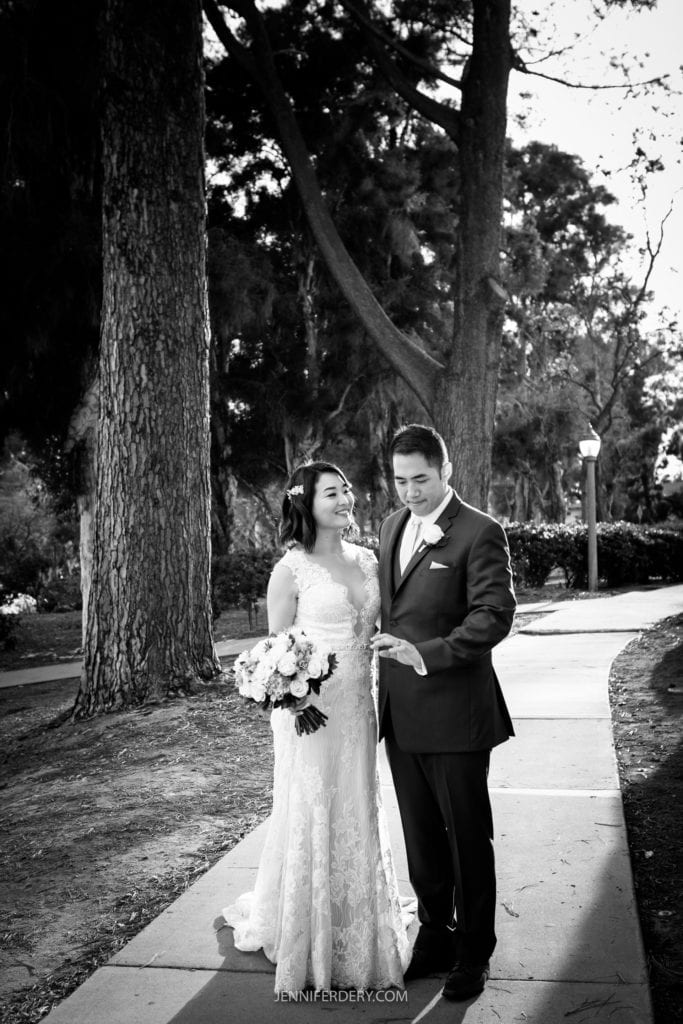 A black-and-white photo of a bride and groom on their wedding day in the Japanese Friendship Garden. The bride, holding a bouquet, is smiling and looking at the groom. The groom, dressed in a suit, is gesturing with his hand while looking at the bride. They are standing on a tree-lined walkway.