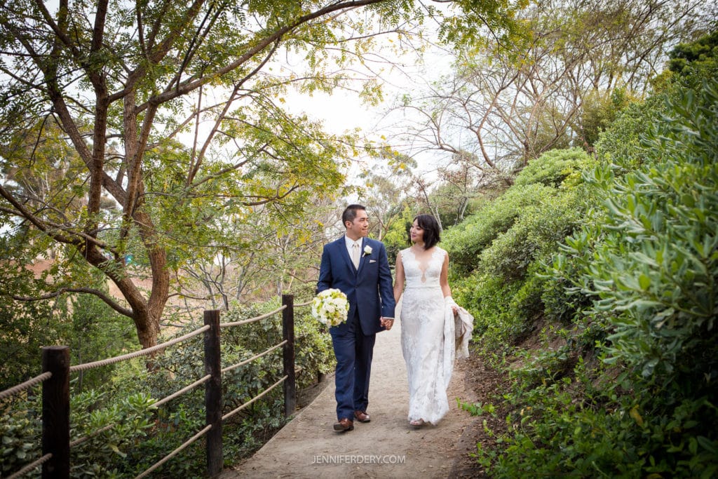A couple in wedding attire walks hand-in-hand along a tree-lined path at the Japanese Friendship Garden. The groom wears a navy blue suit, and the bride wears a lace gown, holding a bouquet. Green foliage surrounds them as they smile at each other, creating a serene and romantic scene.