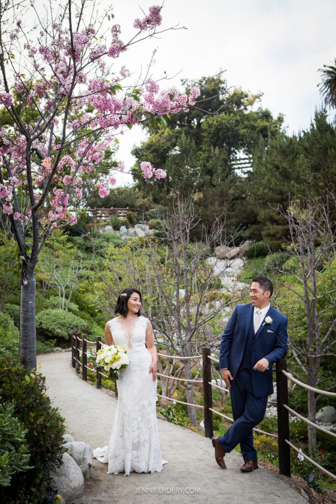 A bride in a white lace gown holds a bouquet and leans against a wooden railing, while a groom in a blue suit stands with his back against the railing, smiling at her. They are outdoors in the lush Japanese Friendship Garden, adorned with blooming cherry blossoms.
