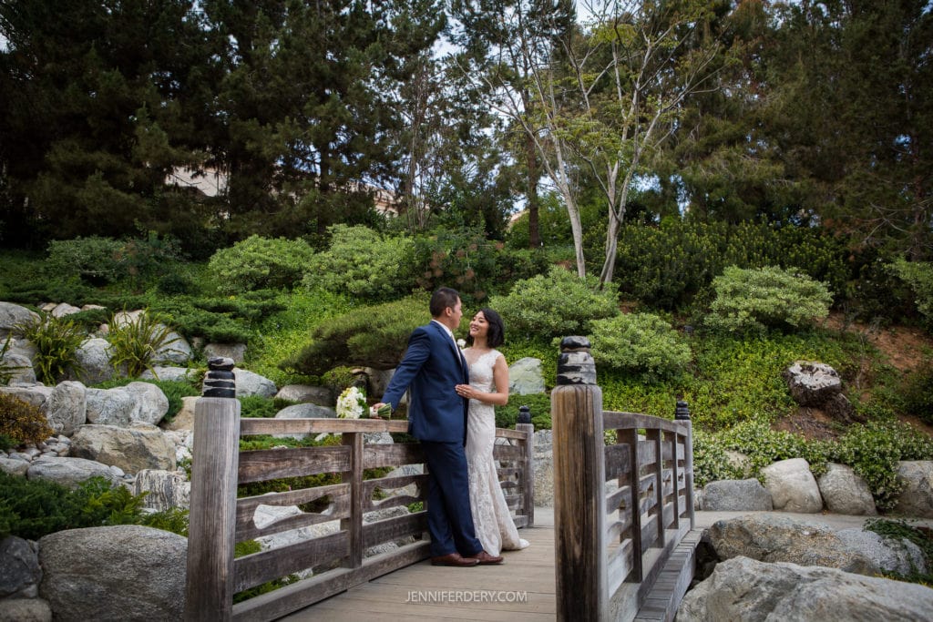 A couple dressed in formal attire stand on a wooden bridge in a lush, green garden at the Japanese Friendship Garden. The man, wearing a blue suit, gazes at the woman in a white dress, who is looking back at him with a smile. Trees and shrubs surround them, creating a picturesque wedding background.
