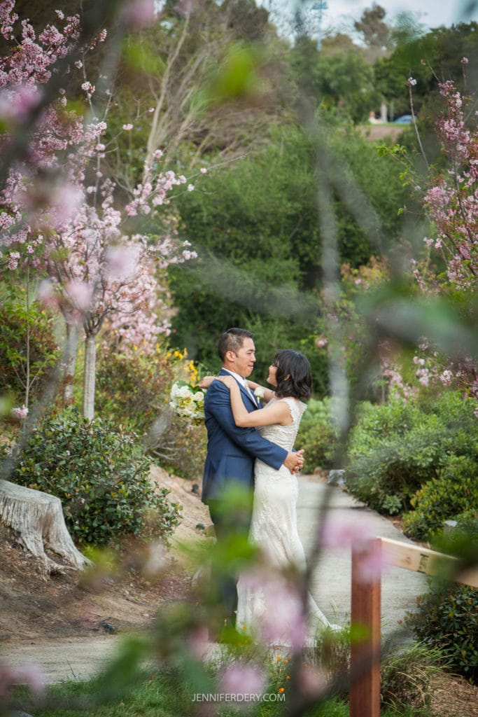 A couple embraces on a pathway surrounded by lush greenery and pink blooming trees at their Japanese Friendship Garden wedding. The bride is wearing a white dress, and the groom is in a blue suit. The scene captures a romantic moment in this serene garden setting.