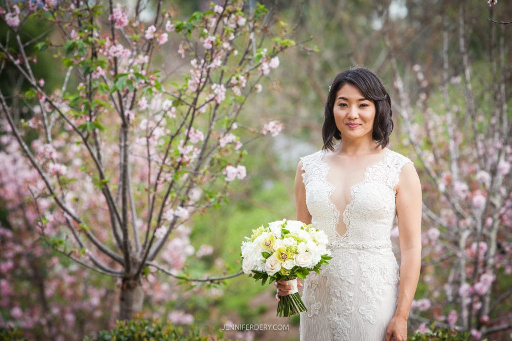 A woman in a white lace wedding dress holds a bouquet of white and yellow flowers. She stands outdoors amid blooming trees and lush greenery at the Japanese Friendship Garden Wedding, with a serene expression on her face.