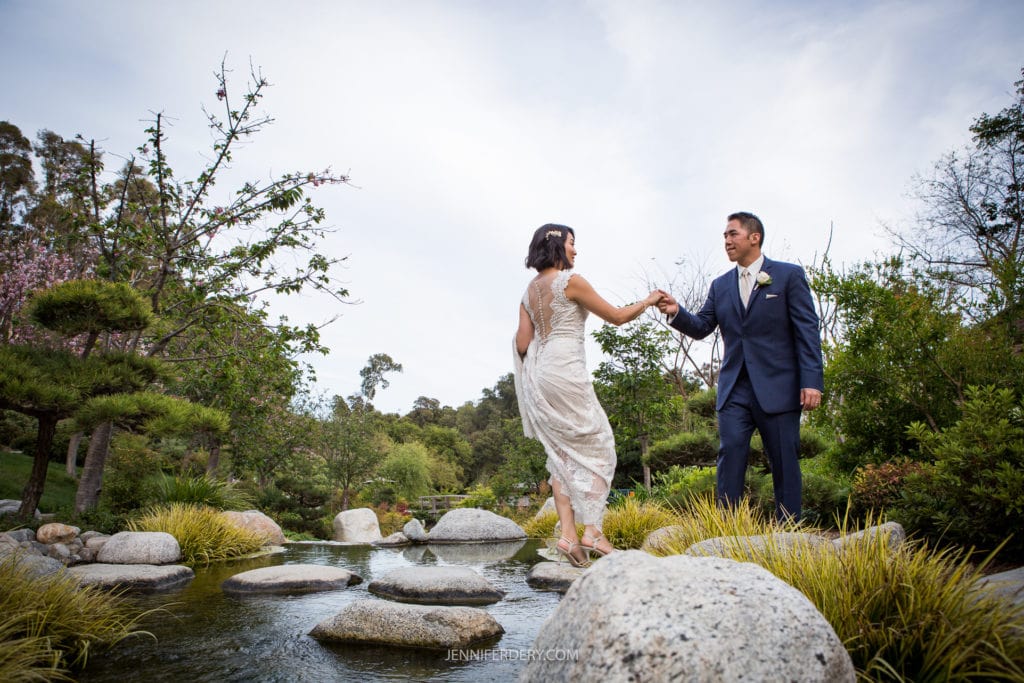 A couple dressed in wedding attire stands by a serene pond with stones and greenery at the Japanese Friendship Garden. The groom in a navy suit gently holds the hand of the bride in a white dress as they balance on the rocks. The background features lush trees and a calm, overcast sky.