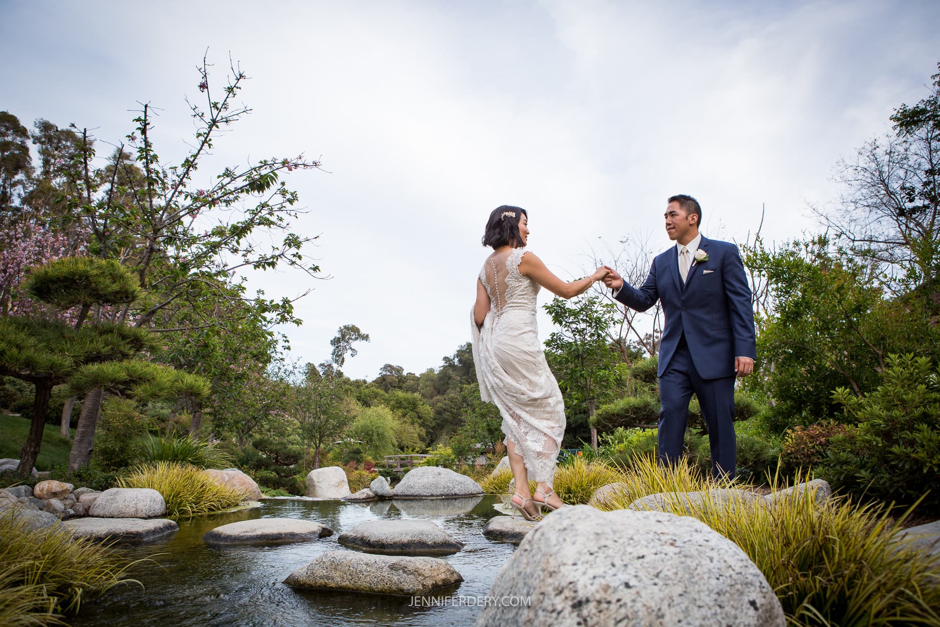 A couple dressed in wedding attire stands by a serene pond with stones and greenery at the Japanese Friendship Garden. The groom in a navy suit gently holds the hand of the bride in a white dress as they balance on the rocks. The background features lush trees and a calm, overcast sky.