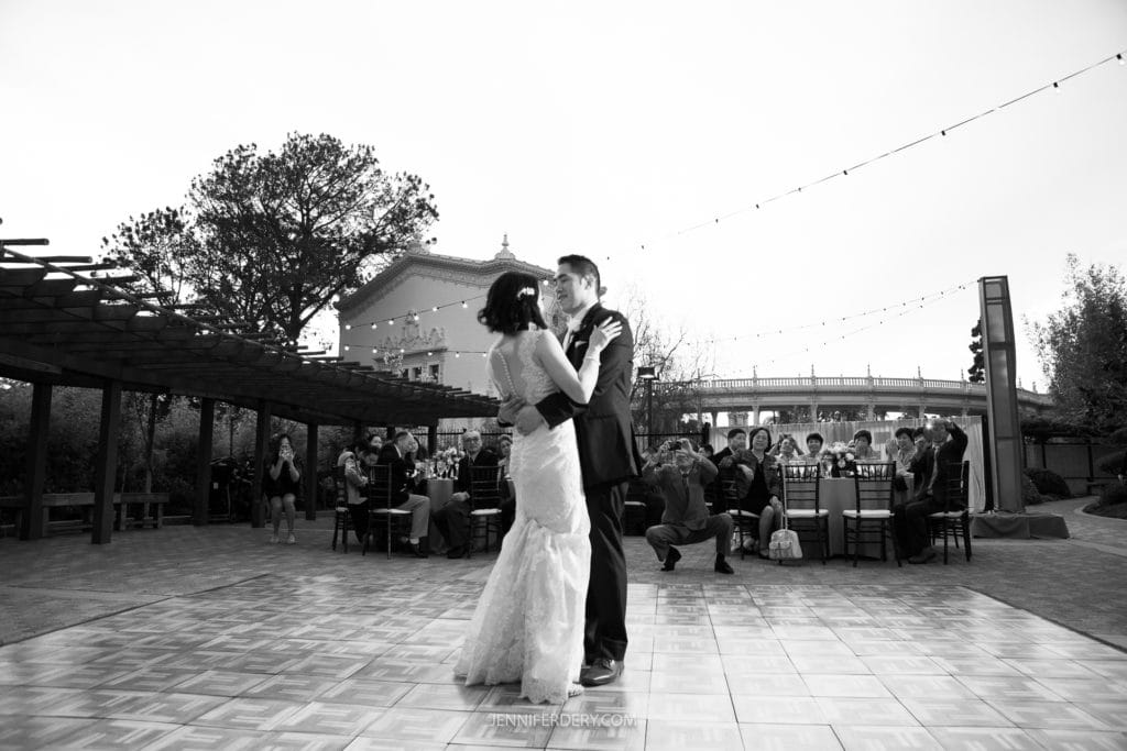 A couple shares a romantic first dance at their Japanese Friendship Garden wedding. They are surrounded by seated guests, some of whom are capturing the moment with their phones. String lights and a building are visible in the background, creating a heartfelt and joyous scene.