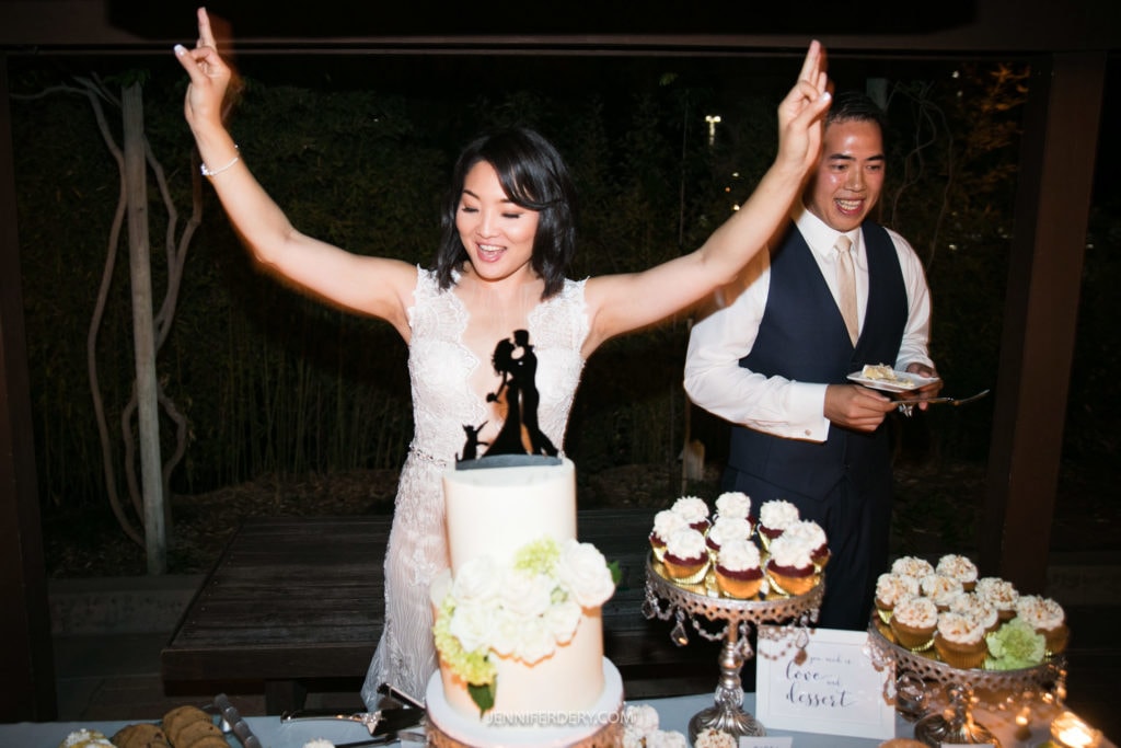 At their wedding reception at the Japanese Friendship Garden, the bride joyfully raises her arms behind a white multi-layered cake topped with a silhouette of a couple. The groom, wearing a vest and tie, smiles while holding a plate of cake. Various desserts surround them.