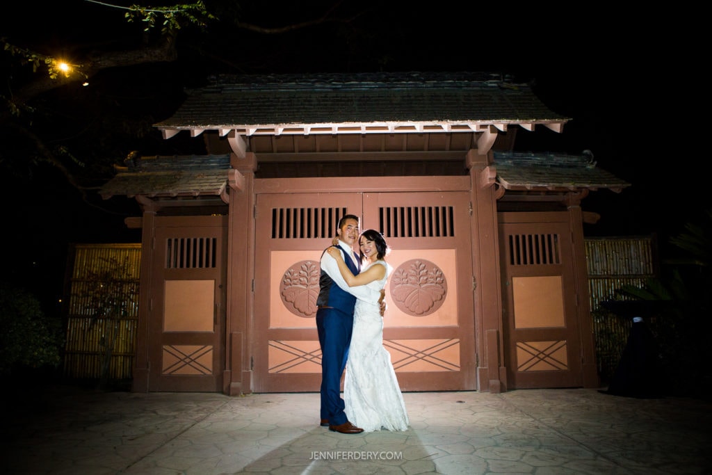 A bride and groom stand together, smiling and embracing, in front of an illuminated wooden gate at night. The bride wears a white wedding dress, and the groom is in a blue suit. Lush greenery and a tree branch frame the scene, adding a touch of magic to this Japanese Friendship Garden wedding.