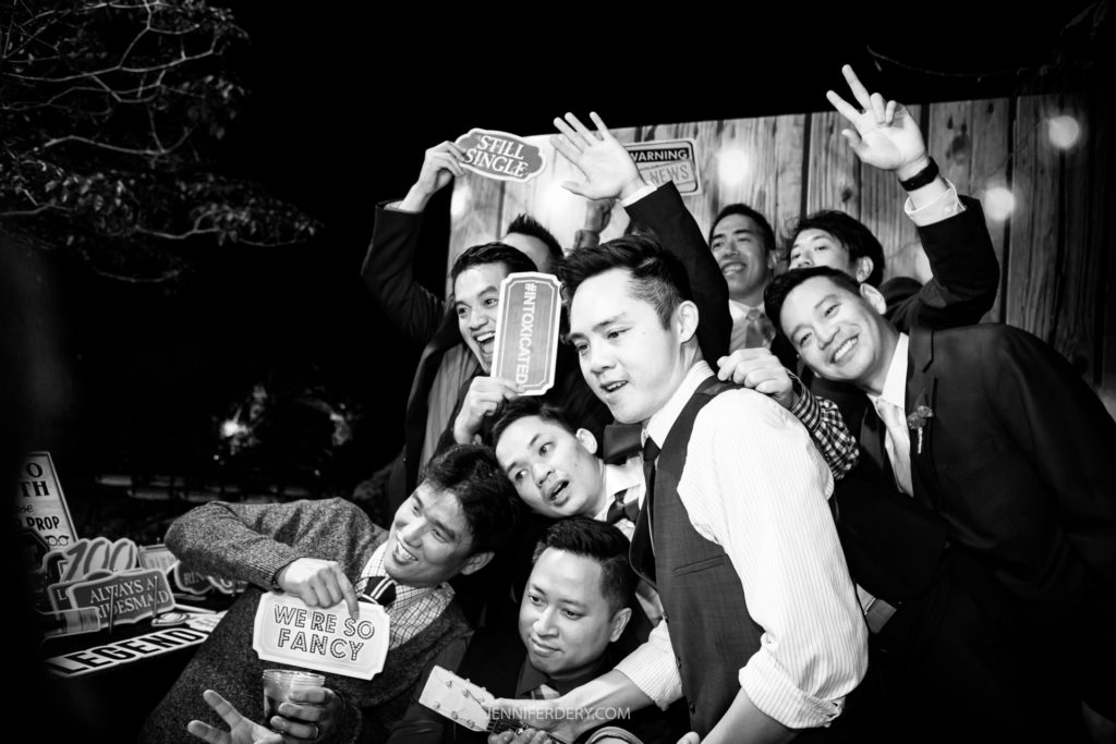 A group of men crowded together in a fun, energetic pose, some holding up humorous signs like "We're So Fancy," "Still Single," and "I'm Here for the Party." Dressed in formal attire, they seem to be celebrating at a Japanese Friendship Garden wedding, with the background dimly lit.