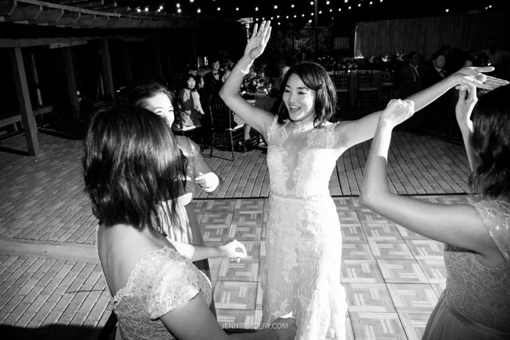 Black and white photo of a joyful bride dancing with three bridesmaids at an outdoor nighttime Japanese Friendship Garden wedding reception. The bride is in a lace gown, her arms raised, and everyone is smiling. String lights are visible in the background.