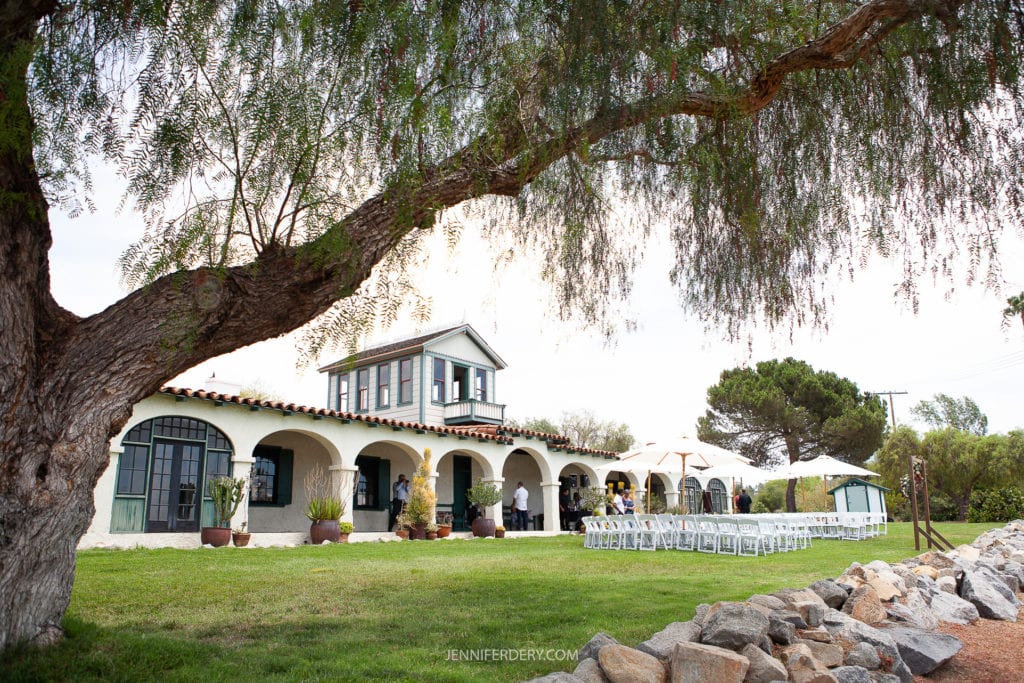 at Rancho Guajome Adobe A picturesque outdoor wedding setup under a large tree, with a charming house featuring arched windows and a balcony in the background. White chairs are arranged in rows, facing a wedding arch. The lawn is surrounded by lush greenery and rock landscaping.