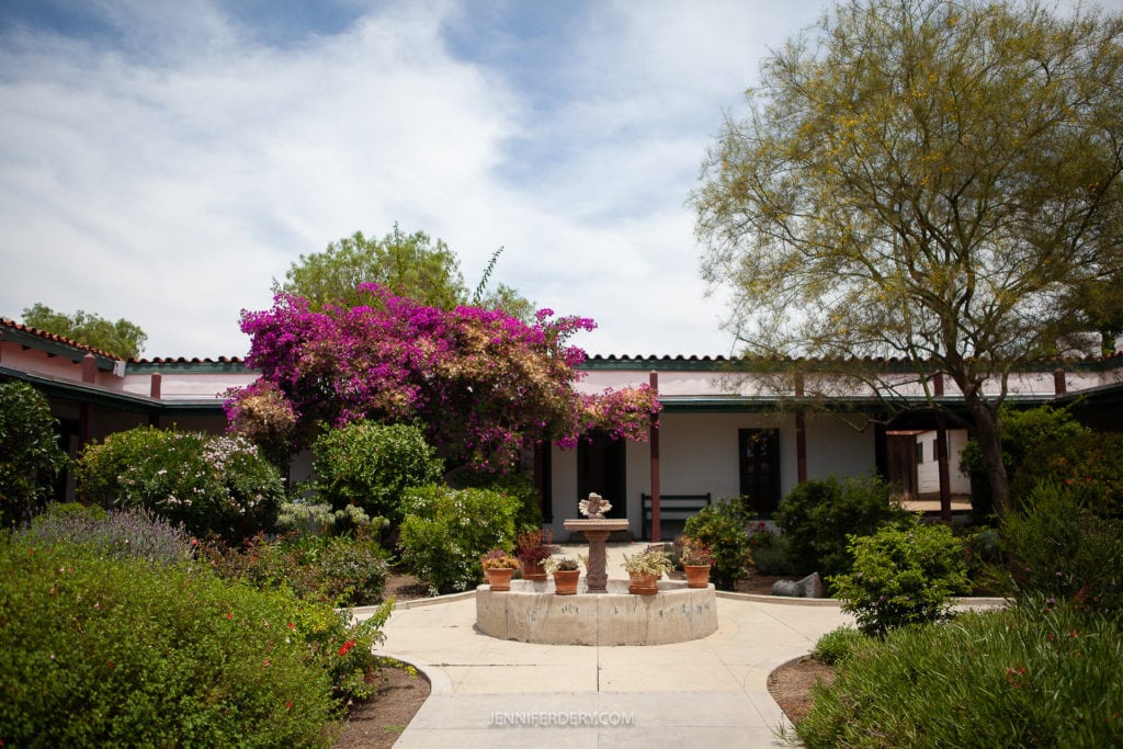A courtyard garden with a central circular stone fountain surrounded by potted plants. Lush greenery and vibrant pink bougainvillea flowers fill the scene. A one-story building with white walls and tiled roof stands in the background under a partly cloudy sky.