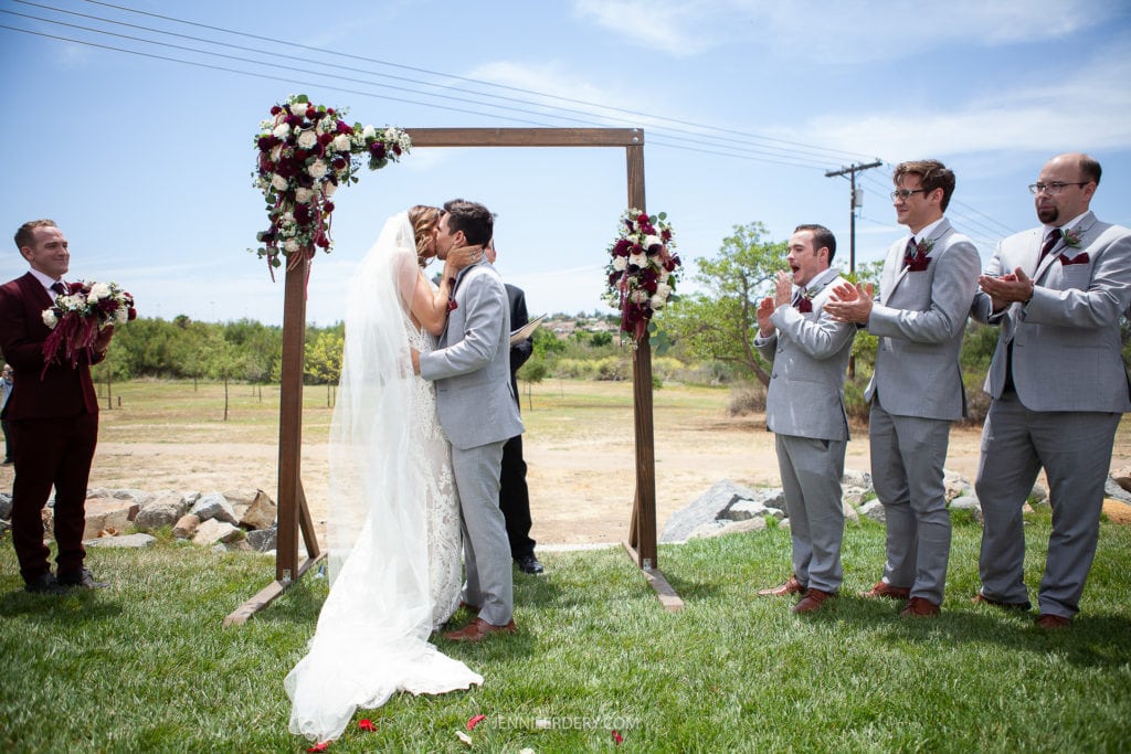 A bride and groom share a kiss under a wooden arch adorned with flowers at an outdoor wedding ceremony at Rancho Guajome Adobe. Two bridesmaids in matching red dresses are on the left holding bouquets, and four groomsmen in gray suits are on the right clapping and smiling.