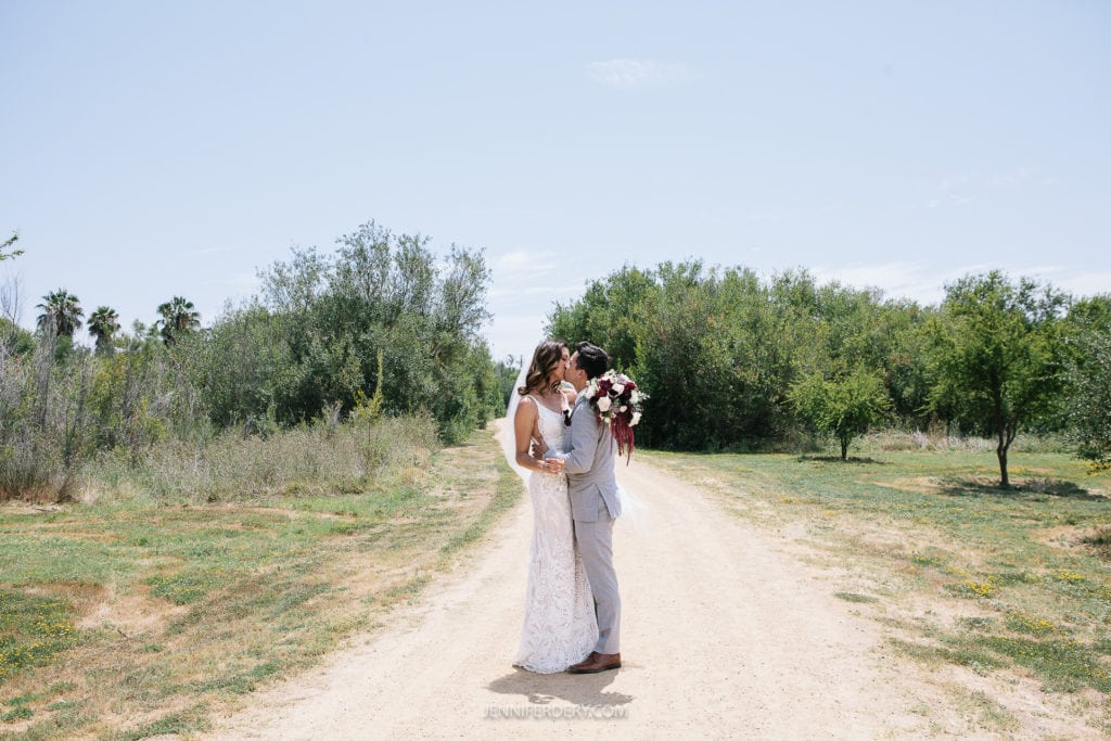 at Rancho Guajome Adobe A couple dressed in wedding attire shares a kiss on a country road surrounded by lush green trees and foliage. The bride is holding a bouquet of flowers and wearing a long, white gown, while the groom is in a light-colored suit. The sky above is clear and blue.