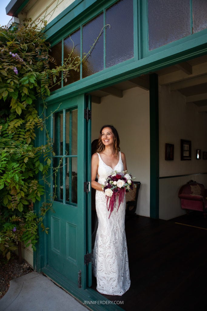A woman in a white lace wedding dress stands in an open doorway under green wooden framing at Rancho Guajome Adobe. She holds a bouquet of flowers, smiling and gazing to the side. Lush greenery and ivy frame the top of the doorway, and faint details of an interior room are visible.