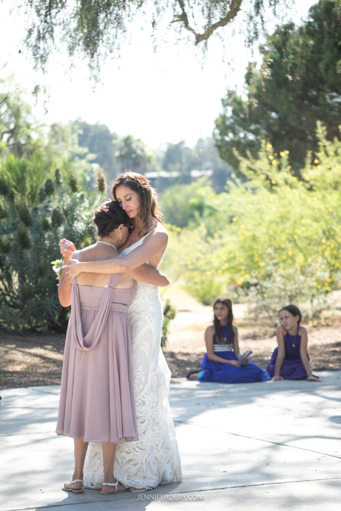 A bride in a white lace dress embraces her mother in a lavender dress outdoors, with greenery in the background. Two young girls in purple dresses sit on the ground in the background. The scene is sunlit and serene.