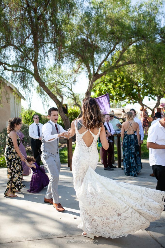 A wedding couple, wearing wedding attire, dances their 1st dance energetically outdoors. The bride's dress has a flowing train and intricate lace details. Friends and family, dressed in semi-formal attire, watch and celebrate under the shade of tall trees in a sunlit setting.